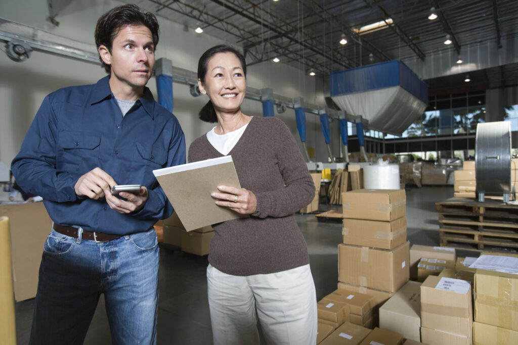 Two team leaders stand next to each other in a warehouse. They appear to be coordinating and planning tasks. One of them is holding a folder or tablet, while the other is making notes in a notebook. In the background, shelves with inventory and logistical equipment are visible